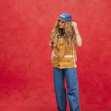 Female model with long-blonde hair wearing 'The Sopranos' Satriales (Pruh-Zhoot) with flowy jeans, brown loafers, and a denim hat with a brown suede brim that says 'Sopranos,' with one hand up towards her hair and the other behind her back, shot on a red background.