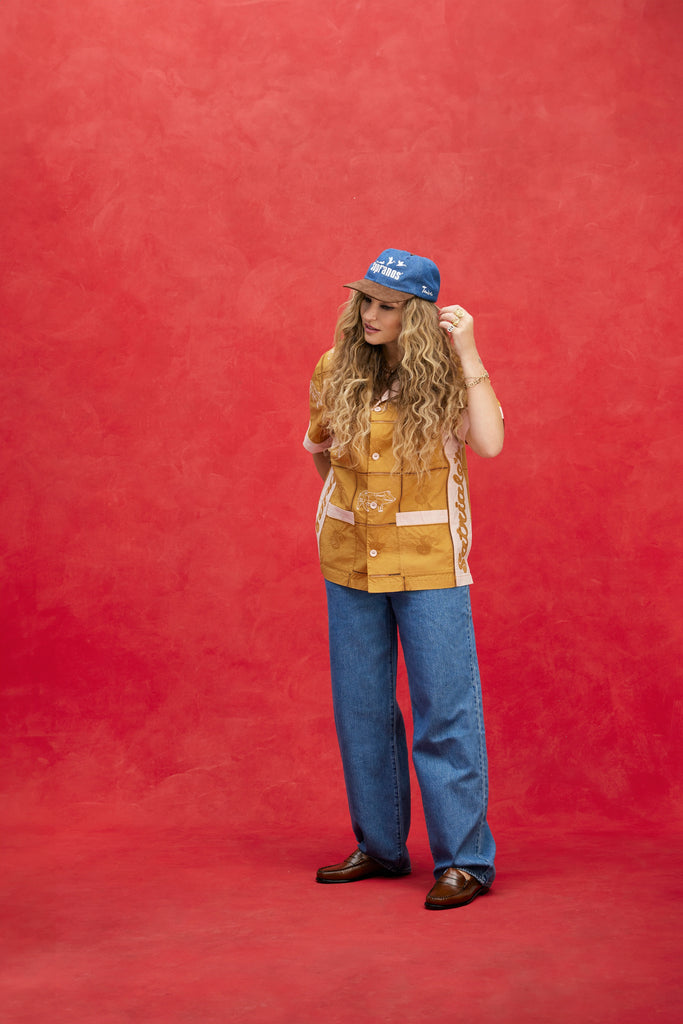 Female model with long-blonde hair wearing 'The Sopranos' Satriales (Pruh-Zhoot) with flowy jeans, brown loafers, and a denim hat with a brown suede brim that says 'Sopranos,' with one hand up towards her hair and the other behind her back, shot on a red background.