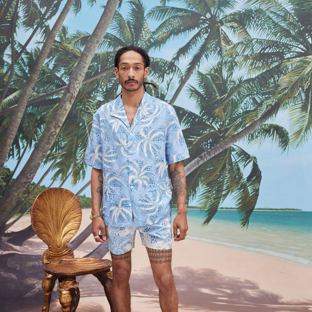 Male model with dreadlocks wearing Palm Fronds Blue with matching Palm Fronds Blue shorts, blue slippers, standing against a background of palm trees and a sunny beach.