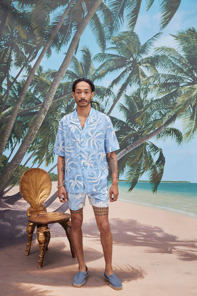 Male model with dreadlocks wearing Palm Fronds Blue with matching Palm Fronds Blue shorts, blue slippers, standing against a background of palm trees and a sunny beach.