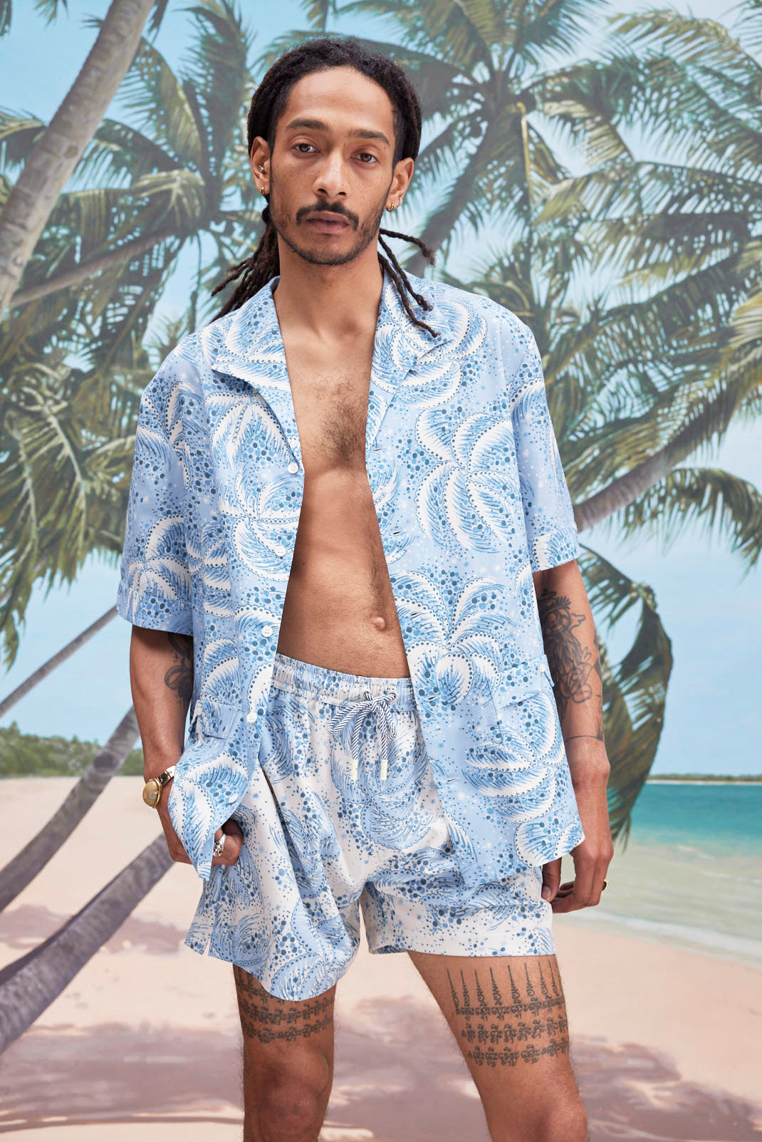 Male model with dreadlocks wearing an unbuttoned 'Palm Fronds (blue)' alongside matching 'Palm Fronds (blue) Swim Trunks,' all shot on a background with palm trees, sun, and a beach. 