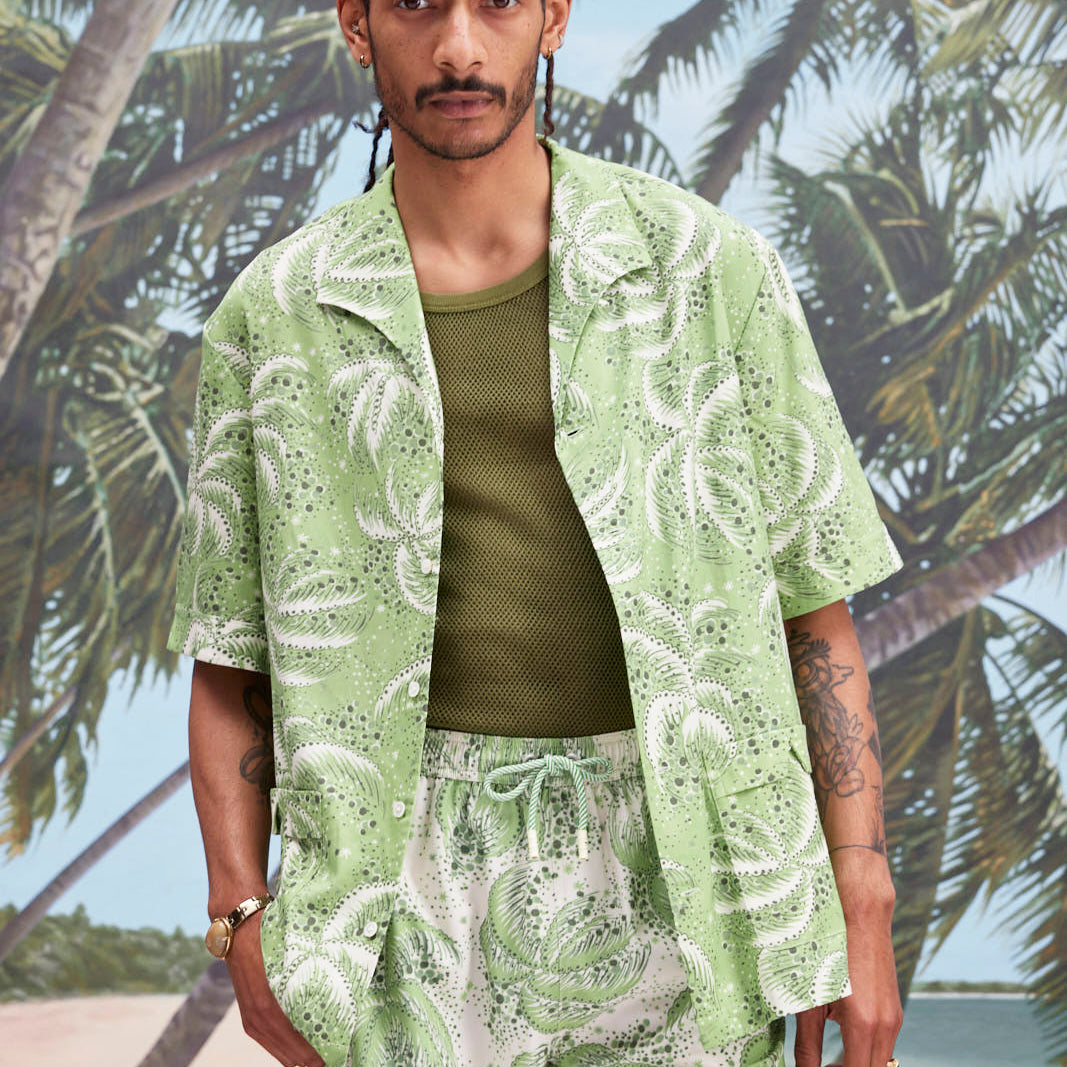 Male model with dreadlocks wearing an unbuttoned 'Palm Fronds (green)' with a dark green tank underneath, and matching 'Palm Fronds (green)' shorts, all shot on a background with palm trees, sun, and a beach.