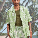 Male model with dreadlocks wearing an unbuttoned 'Palm Fronds (green)' with a dark green tank underneath, and matching 'Palm Fronds (green)' shorts, all shot on a background with palm trees, sun, and a beach.