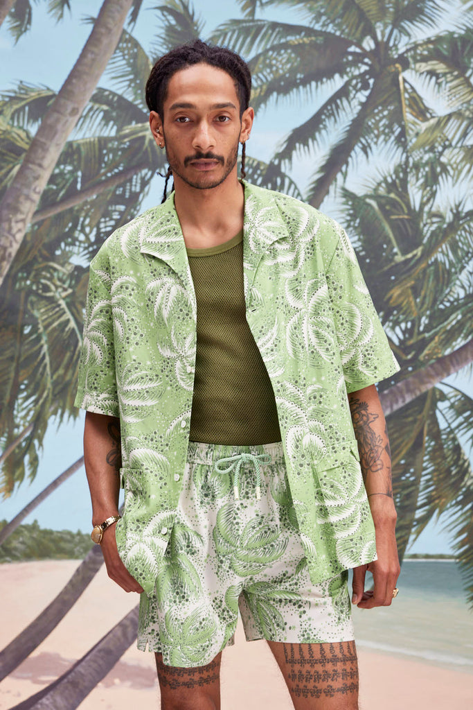 Male model with dreadlocks wearing an unbuttoned 'Palm Fronds (green)' with a dark green tank underneath, and matching 'Palm Fronds (green)' shorts, all shot on a background with palm trees, sun, and a beach.