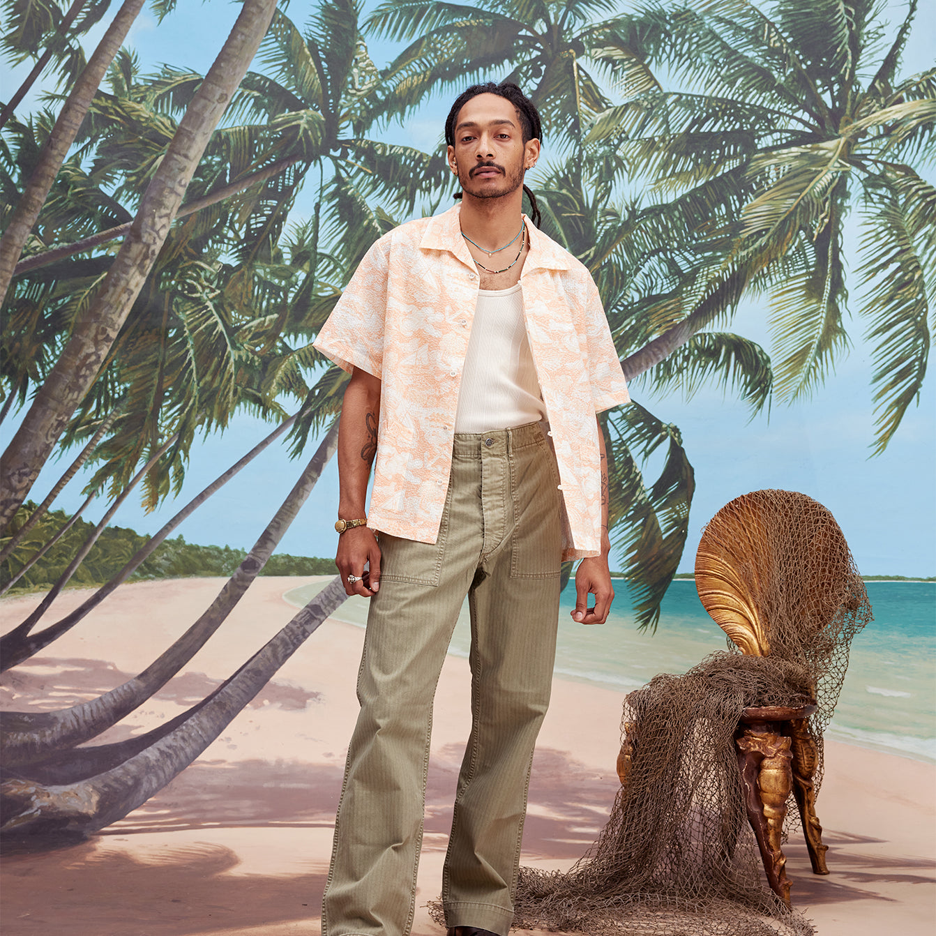 Male model with dreadlocks wearing the 'Voyager Shirt' in coral seersucker unbuttoned with a white tank top underneath and light green pants, and dark brown sandals,  shot on a background with palm trees, blue skies, and a beach.