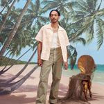Male model with dreadlocks wearing the 'Voyager Shirt' in coral seersucker unbuttoned with a white tank top underneath and light green pants, and dark brown sandals,  shot on a background with palm trees, blue skies, and a beach.