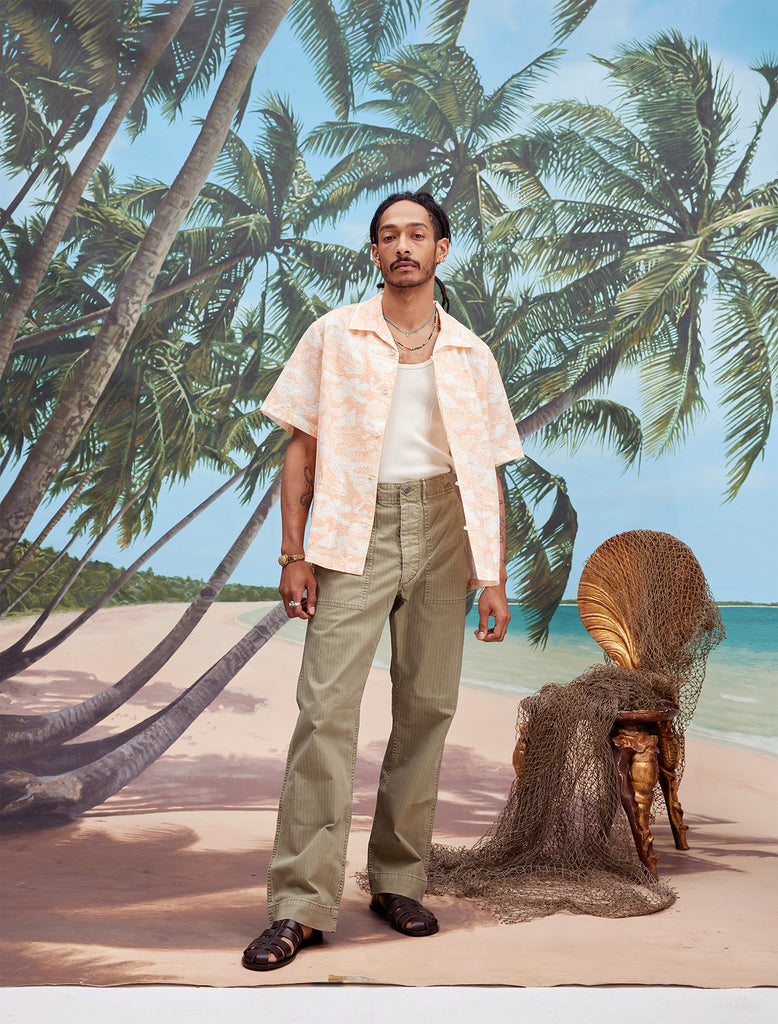 Male model with dreadlocks wearing the 'Voyager Shirt' in coral seersucker unbuttoned with a white tank top underneath and light green pants, and dark brown sandals,  shot on a background with palm trees, blue skies, and a beach.