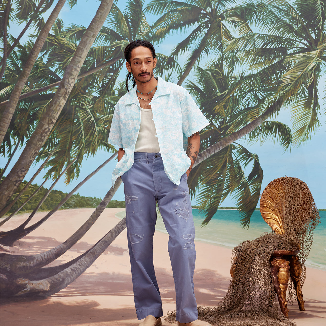 Male model with dreadlocks wearing the 'Voyager Shirt' in Aqua Seersucker unbuttoned with a white tanktop and indigo jeans with beige espadrilles, shot on a background with a beach, blue skies, and palm trees.
