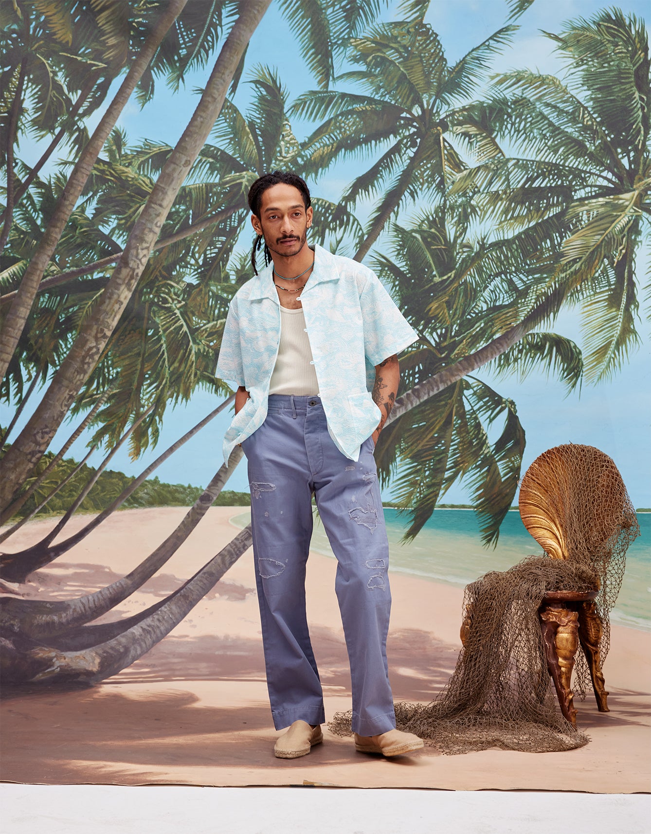 Male model with dreadlocks wearing the 'Voyager Shirt' in Aqua Seersucker unbuttoned with a white tanktop and indigo jeans with beige espadrilles, shot on a background with a beach, blue skies, and palm trees.