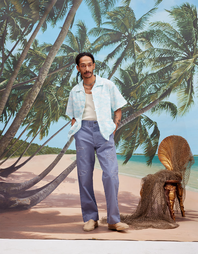 Male model with dreadlocks wearing the 'Voyager Shirt' in Aqua Seersucker unbuttoned with a white tanktop and indigo jeans with beige espadrilles, shot on a background with a beach, blue skies, and palm trees.