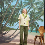 A woman wearing a cream-colored short-sleeve shirt with green trim and a large banana applique on the front, green pants, and a light scarf tied around her neck. 