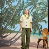 A woman wearing a cream-colored short-sleeve shirt with green trim and a large banana applique on the front, green pants, and a light scarf tied around her neck. 