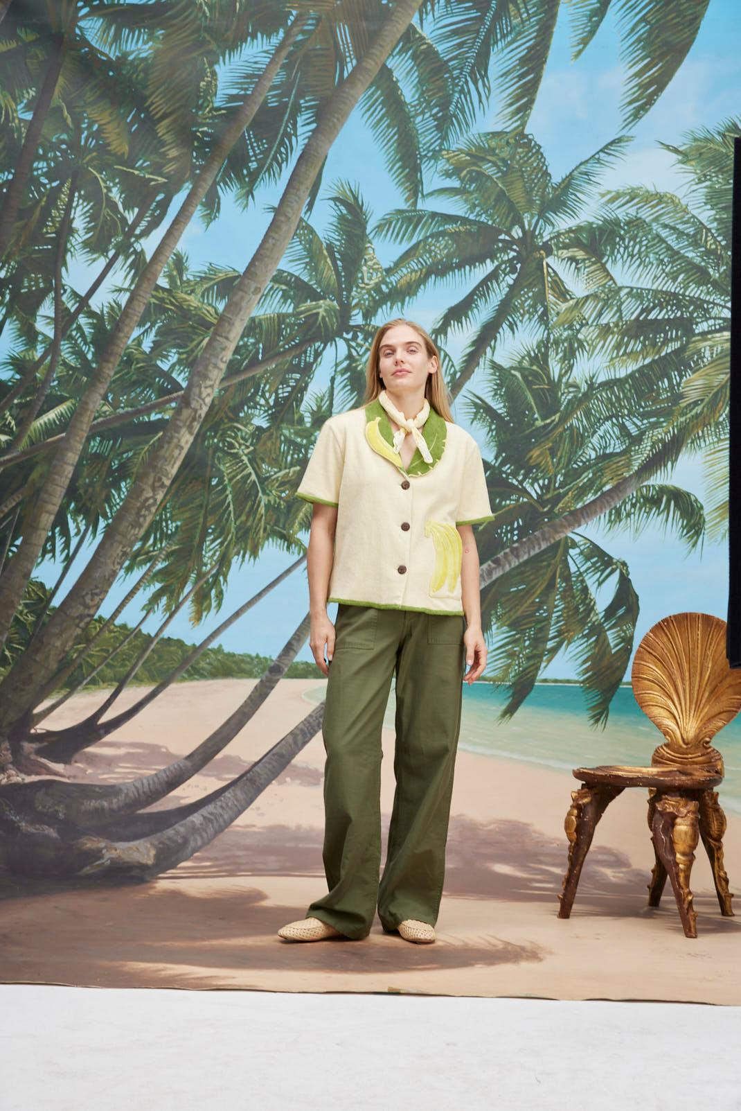 A woman wearing a cream-colored short-sleeve shirt with green trim and a large banana applique on the front, green pants, and a light scarf tied around her neck. 