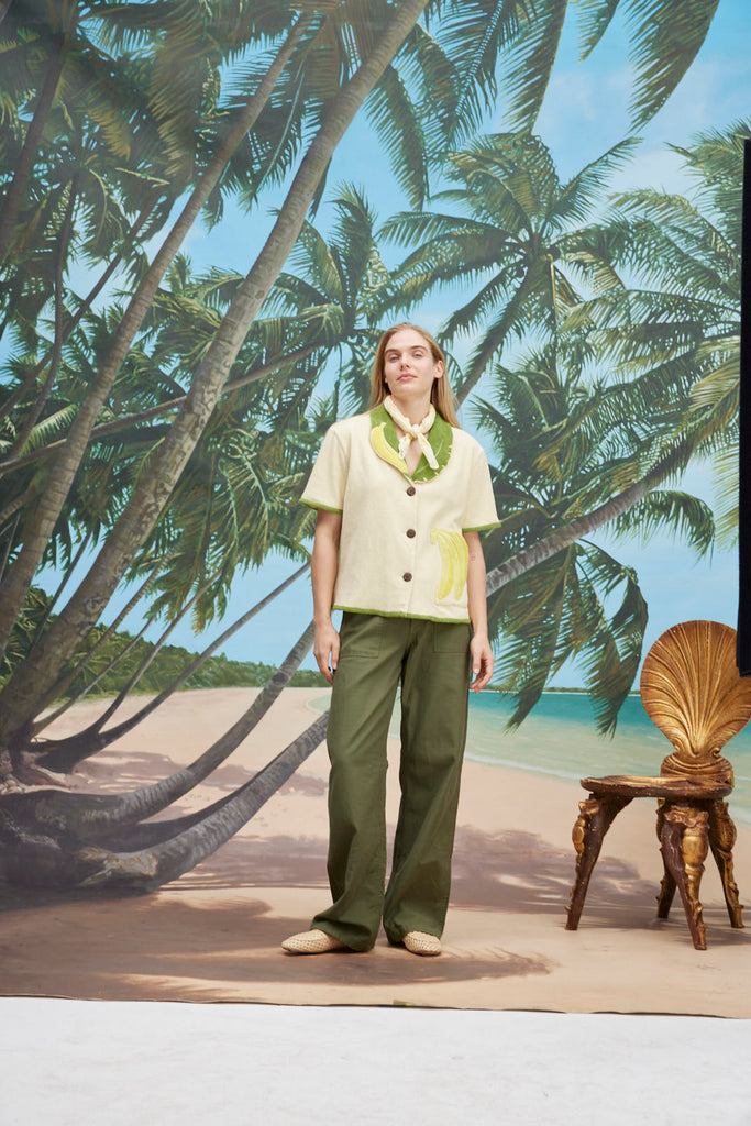 A woman wearing a cream-colored short-sleeve shirt with green trim and a large banana applique on the front, green pants, and a light scarf tied around her neck. 