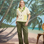 A woman wearing a cream-colored short-sleeve shirt with green trim and a large banana applique on the front, green pants, and a light scarf tied around her neck. 