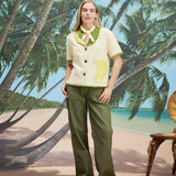 A woman wearing a cream-colored short-sleeve shirt with green trim and a large banana applique on the front, green pants, and a light scarf tied around her neck. 