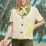 A woman wearing a cream-colored short-sleeve shirt with green trim and a large banana applique on the front, green pants, and a light scarf tied around her neck. 