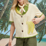 A woman wearing a cream-colored short-sleeve shirt with green trim and a large banana applique on the front, green pants, and a light scarf tied around her neck. She appears to be laughing.