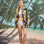 Young female model with blonde long hair wearing unbuttoned "Night Bloom" long-sleeve with a white bathing suit on a background of a beach with leaning palm trees and blue skies.