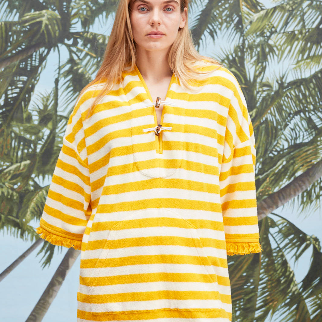 Female model with long blonde hair wearing the 'Saffron Stripe' Beach Towel Top with a white skirt with netting, a beach, palm fronds, and blue skies.