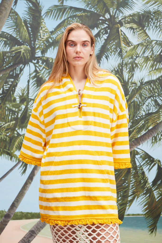 Female model with long blonde hair wearing the 'Saffron Stripe' Beach Towel Top with a white skirt with netting, a beach, palm fronds, and blue skies.