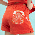 Close-up of orange terry cloth shorts featuring an embroidered design of a white rotary telephone and scallop shell on the back pocket.