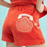 Close-up of orange terry cloth shorts featuring an embroidered design of a white rotary telephone and scallop shell on the back pocket.