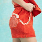 Close-up of orange terry cloth shorts with an embroidered white rotary phone and scallop shell on the back pocket, paired with a matching top featuring a coiled phone cord motif.