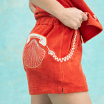 Close-up of orange terry cloth shorts with an embroidered white rotary phone and scallop shell on the back pocket, paired with a matching top featuring a coiled phone cord motif.