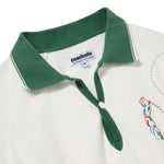 Close-up of a white terry cloth polo shirt with a forest green collar and matching placket. Embroidery on the chest shows a colorful golfer mid-swing, with a dotted line trailing the ball. A woven label inside the collar reads 'tombolo PERFORMANCE INACTIVEWEAR'.