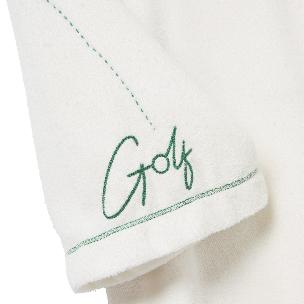Close-up of the left sleeve of a white terry cloth polo shirt. Green embroidery reads 'Golf' in script, with a dotted line extending upward from the lettering. 