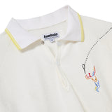 Close-up of a white terry cloth polo shirt with a white collar featuring yellow trim. Embroidery shows a colorful figure swinging a tennis racket, with a dotted line trailing the ball. Two white buttons are visible beneath the collar. A woven label reads 'tombolo PERFORMANCE INACTIVEWEAR'.