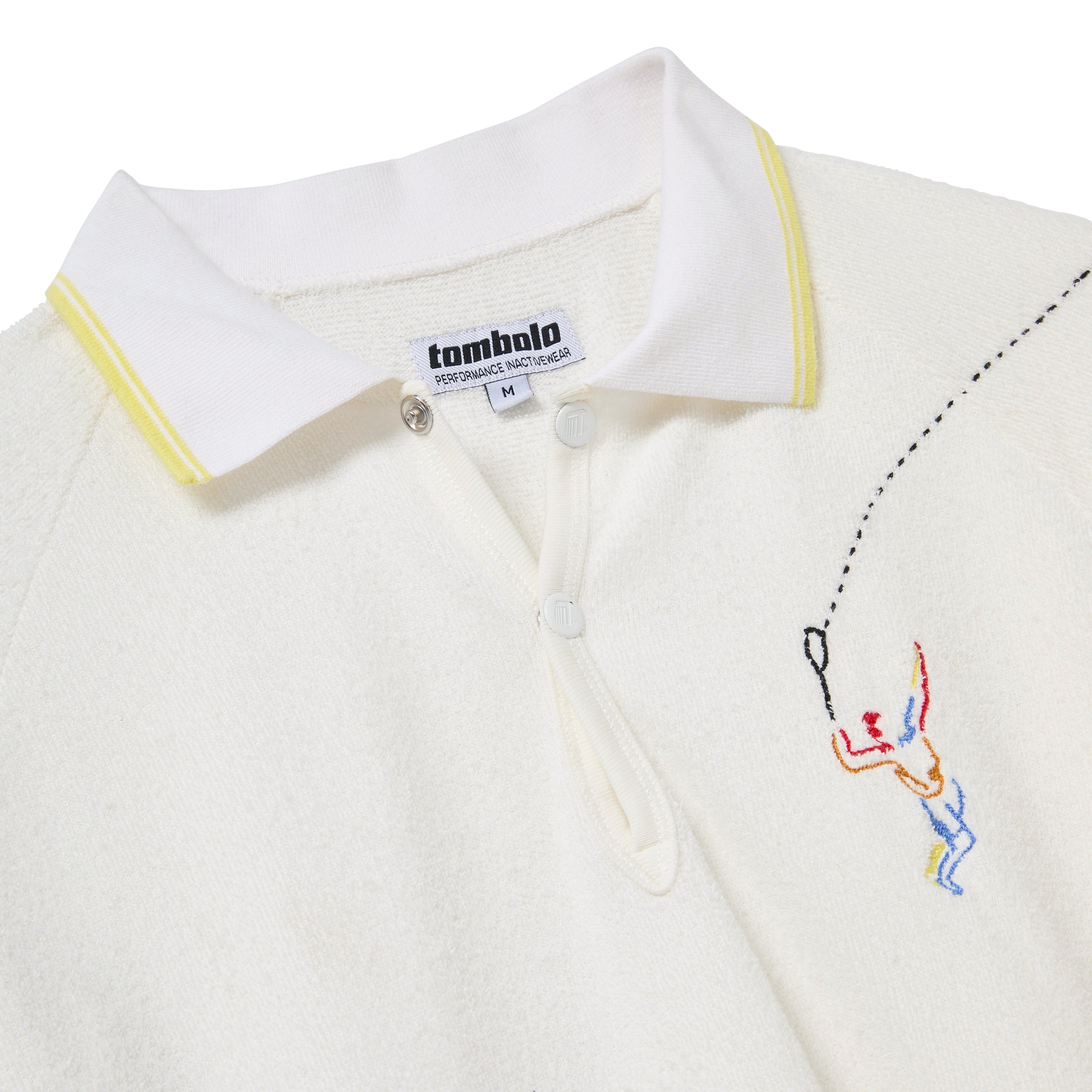 Close-up of a white terry cloth polo shirt with a white collar featuring yellow trim. Embroidery shows a colorful figure swinging a tennis racket, with a dotted line trailing the ball. Two white buttons are visible beneath the collar. A woven label reads 'tombolo PERFORMANCE INACTIVEWEAR'.
