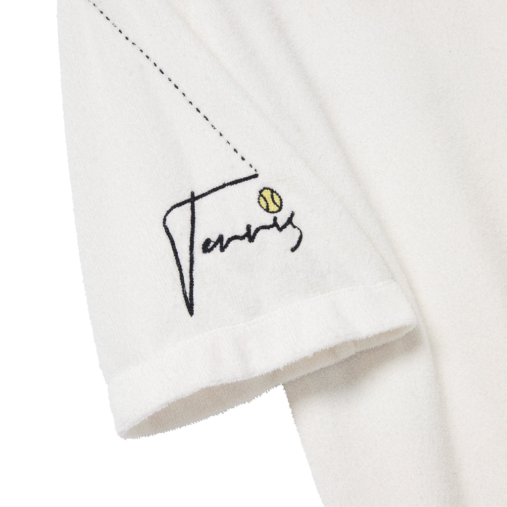 Close-up of the left sleeve of a white terry cloth polo shirt. Embroidery features the word 'Tennis' in black script with a small yellow tennis ball above the letter 'i' and a dotted line trailing upward toward the shoulder.