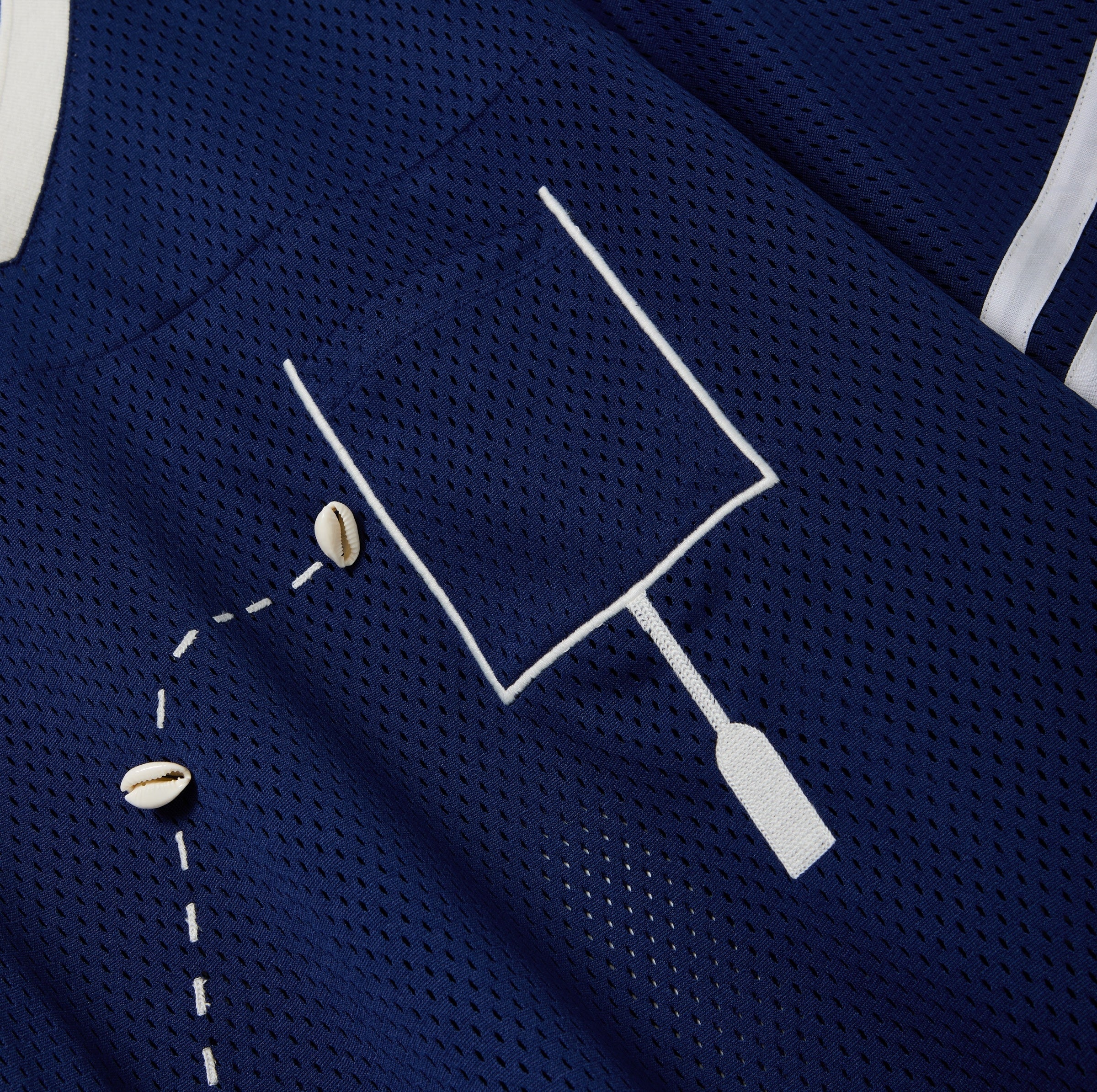 Close-up of a navy blue mesh shirt with a white outlined pocket and cowrie shells arranged to mimic a football in the air.