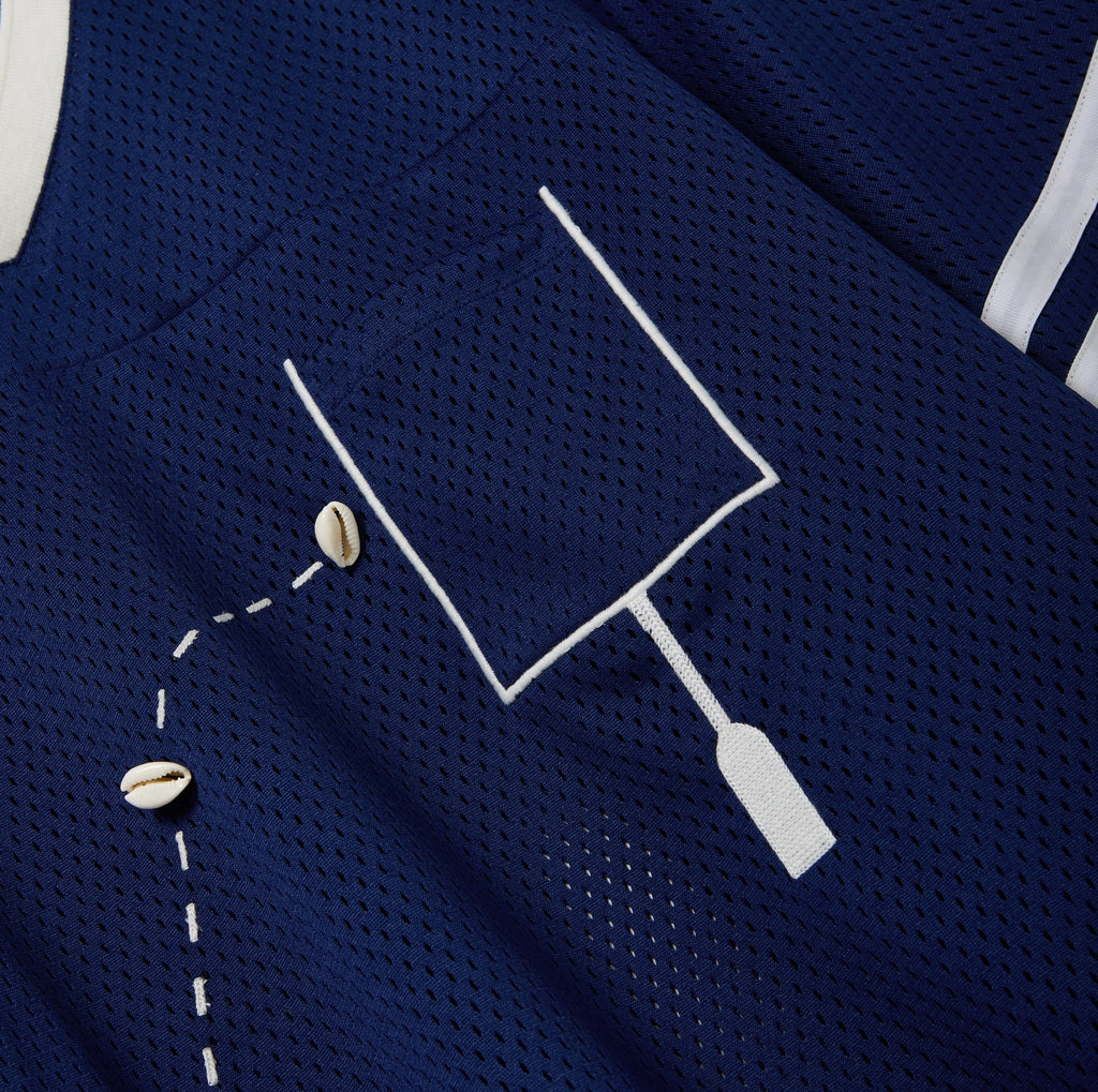 Close-up of a navy blue mesh shirt with a white outlined pocket and cowrie shells arranged to mimic a football in the air.