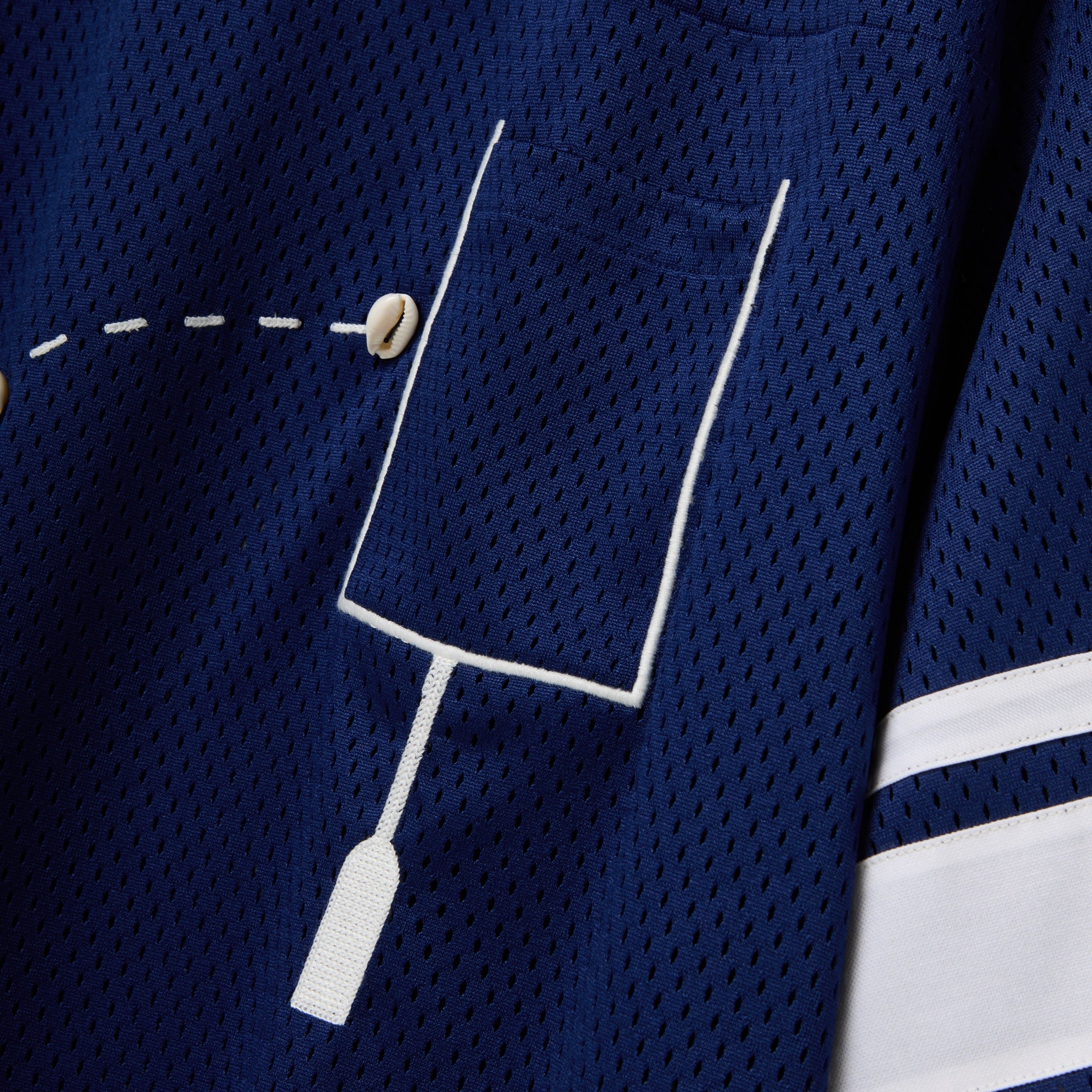 Close-up of a navy blue mesh shirt with a white outlined pocket and cowrie shells arranged to mimic a football in the air.