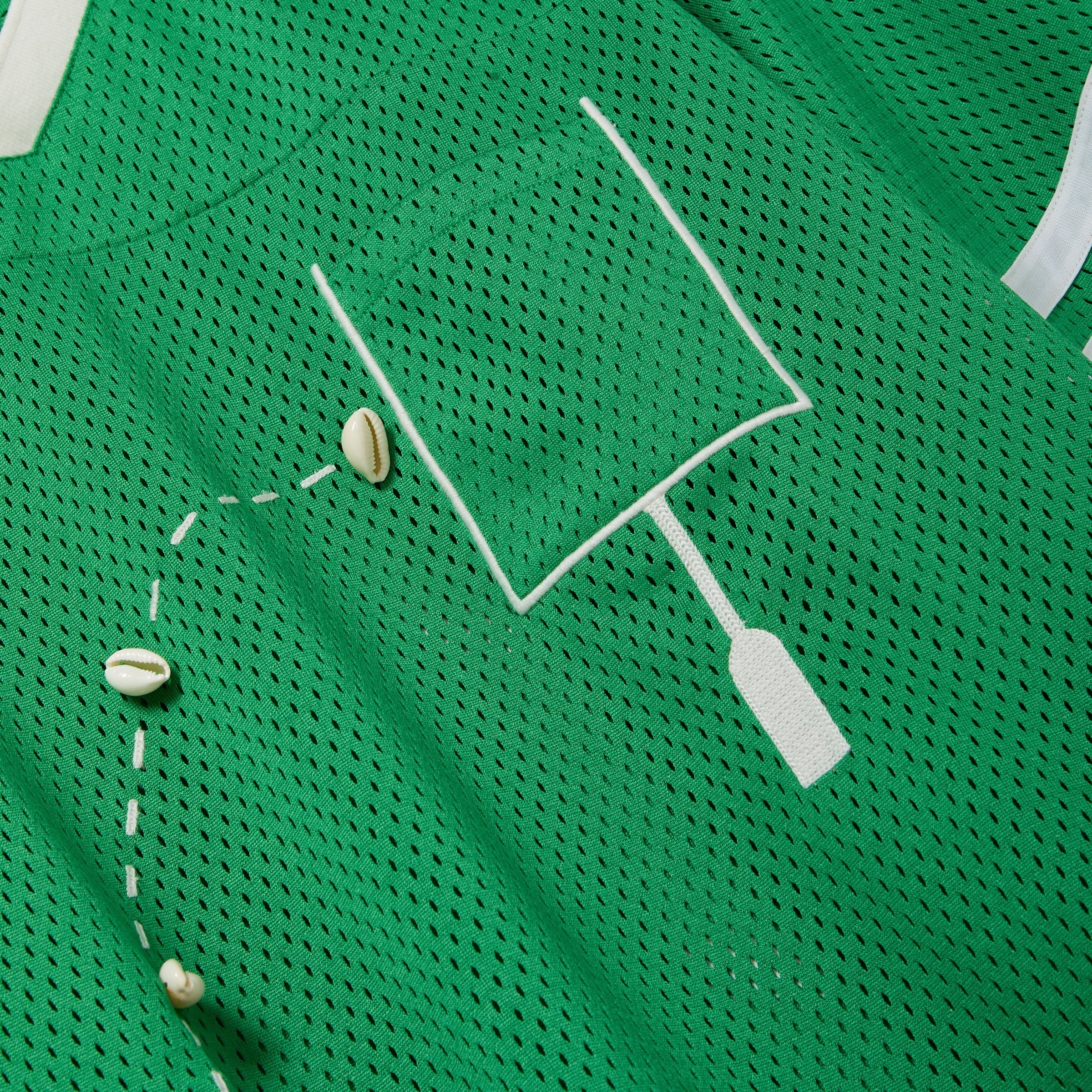 Close-up of a green mesh shirt with a white outlined pocket and cowrie shells arranged to mimic a football graphic.