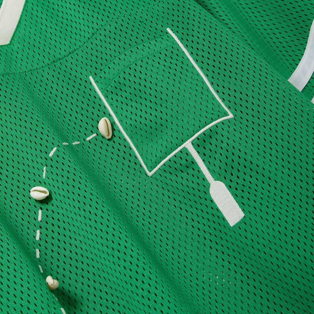 Close-up of a green mesh shirt with a white outlined pocket and cowrie shells arranged to mimic a football graphic.