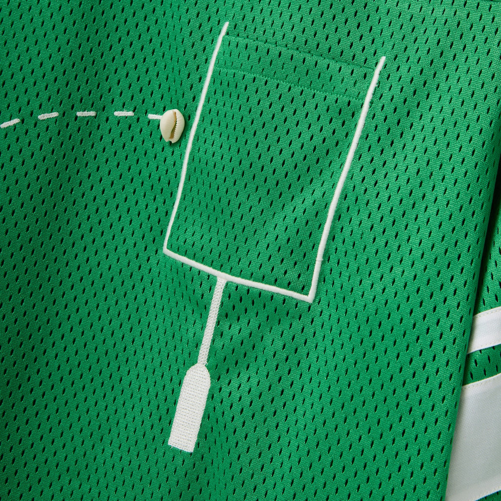 Close-up of a green mesh shirt with a white outlined pocket and cowrie shells arranged to mimic a football graphic.