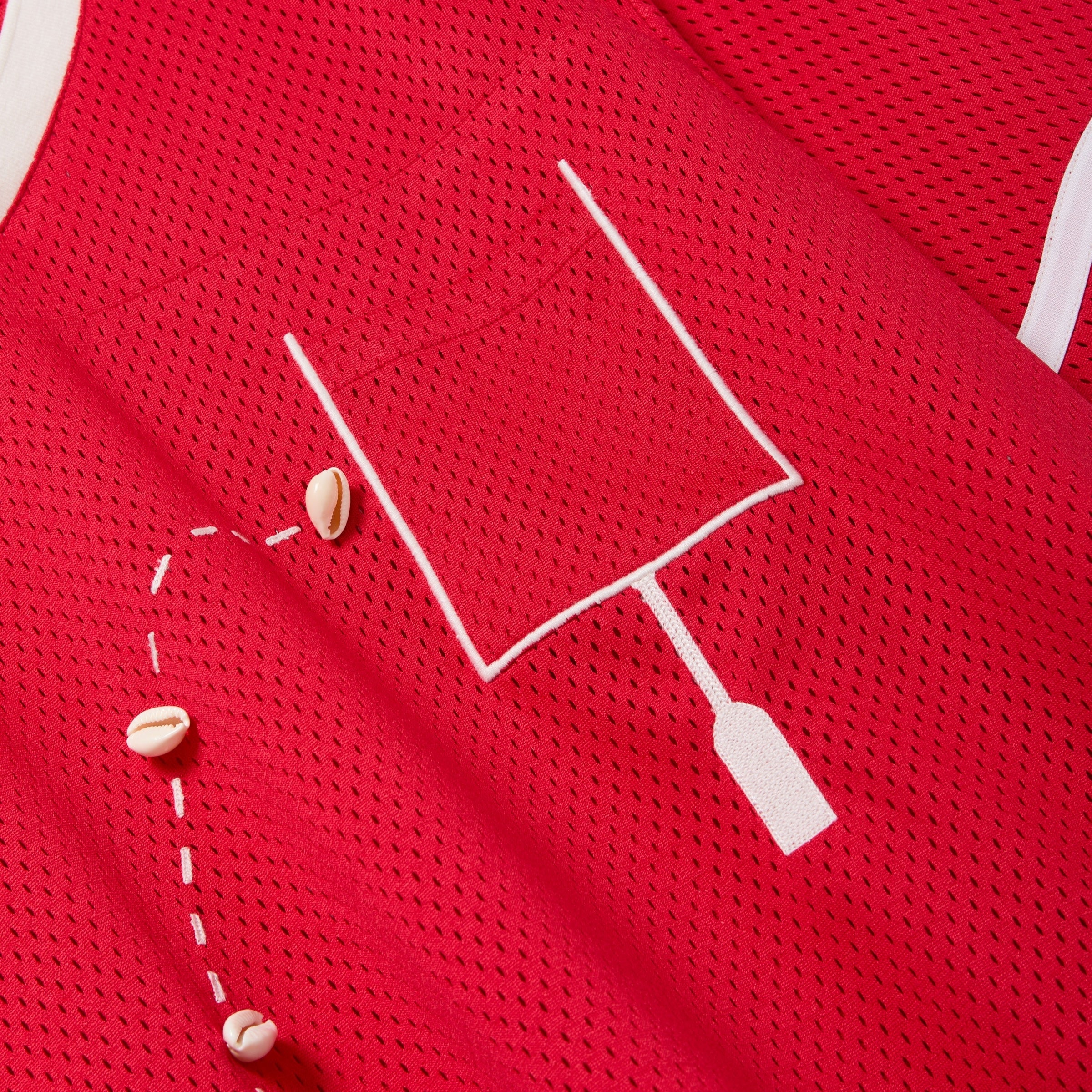 Close-up of a red mesh shirt with a white outlined pocket and cowrie shells arranged to mimic a football graphic.