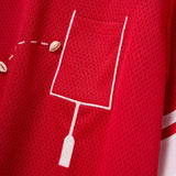Close-up of a red mesh shirt with a white outlined pocket and cowrie shells arranged to mimic a football graphic