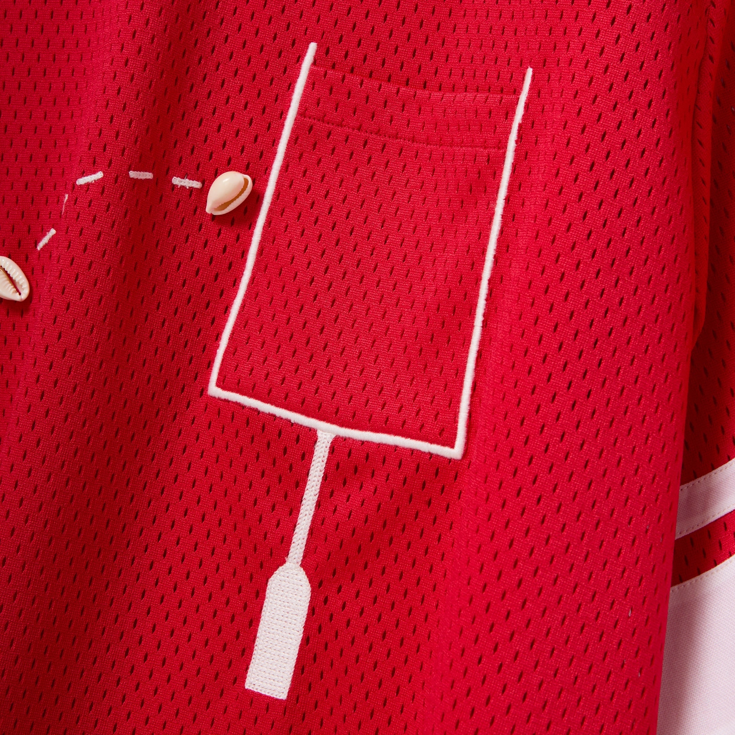 Close-up of a red mesh shirt with a white outlined pocket and cowrie shells arranged to mimic a football graphic