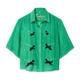 Green cropped cabana shirt with black dog silhouettes across the front, each attached to a white leash motif, with white piping along the collar and sleeves.