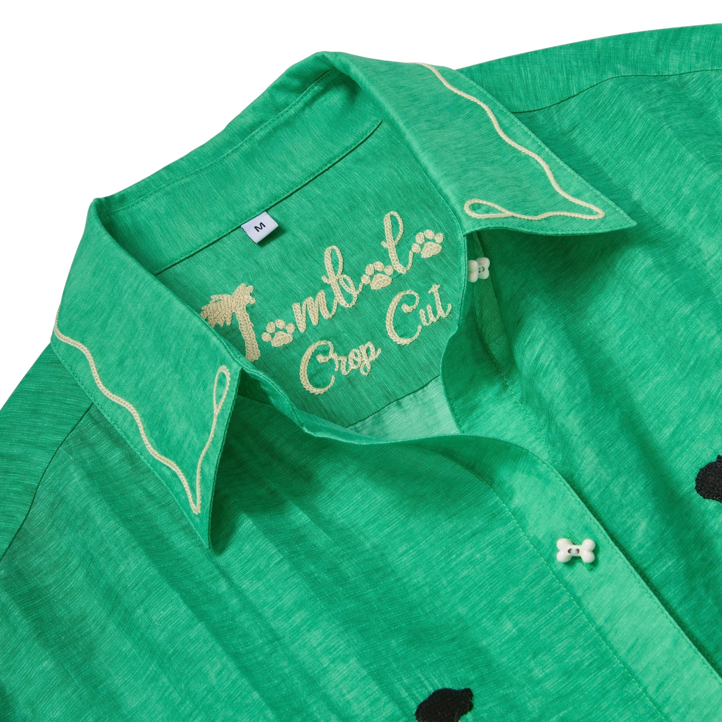 Close-up of a green cropped cabana shirt collar with white leash embroidery and “Tombolo Crop Cut” script stitched inside the neckline.