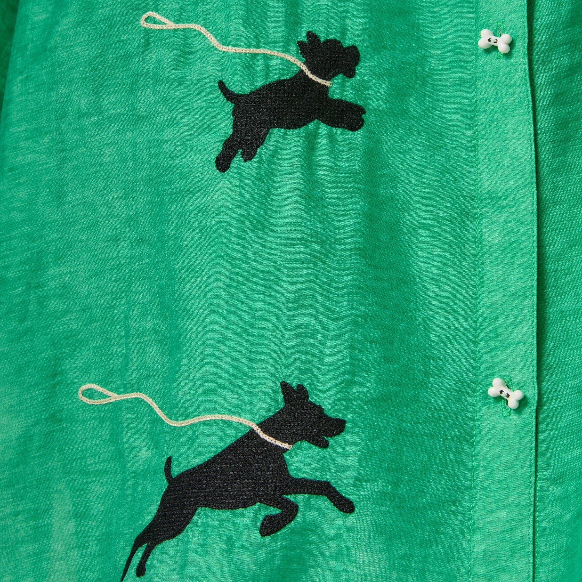 Close-up of a green cabana shirt featuring black embroidered dog silhouettes attached to white leash embroidery and dog bone–shaped buttons.