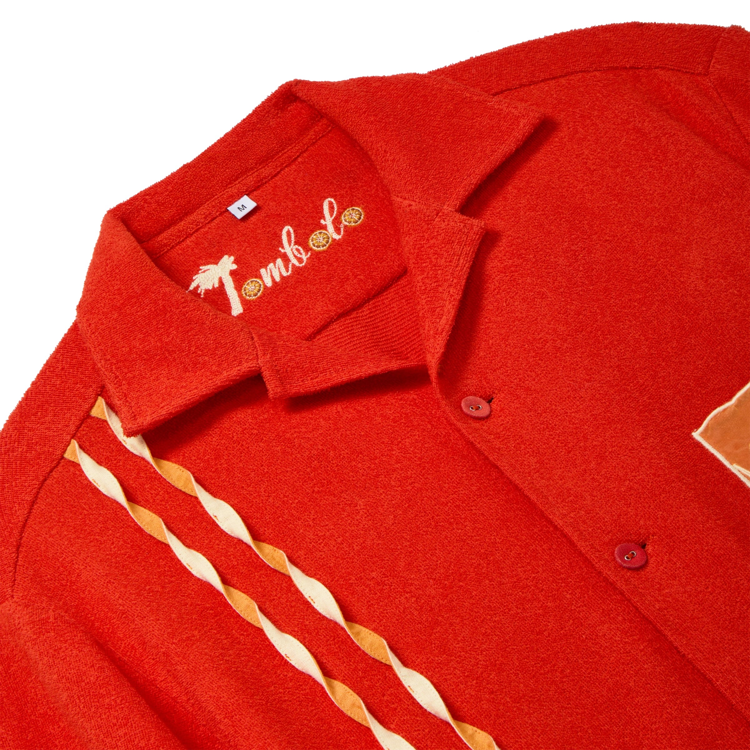 Close-up of a red cabana shirt showing the chain-stitched “Tombolo” logo at the collar and two twisted appliqué stripes running down the front.
