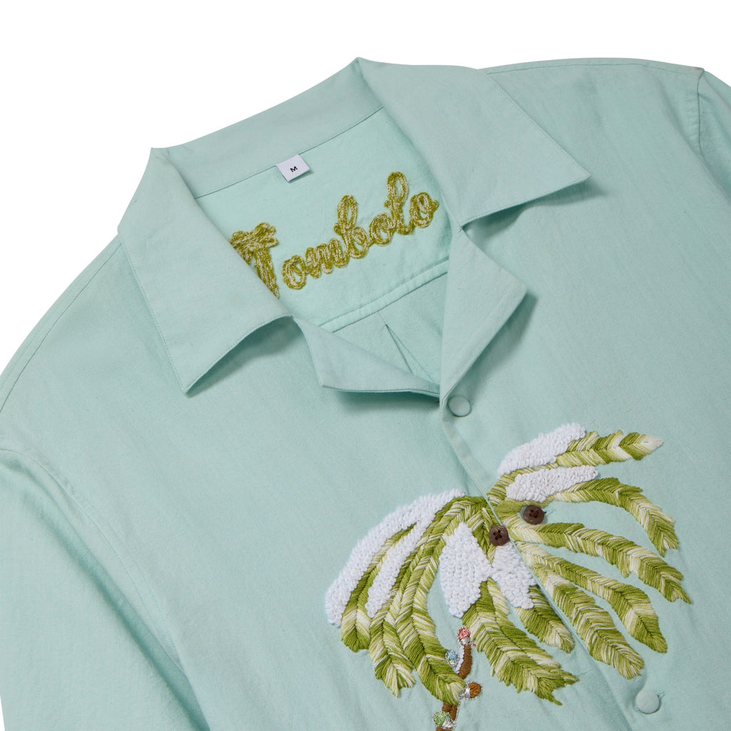 Close-up of a light blue cabana shirt showing the chain-stitched “Tombolo” logo at the collar and embroidered snowy palm tree detailing on the front.