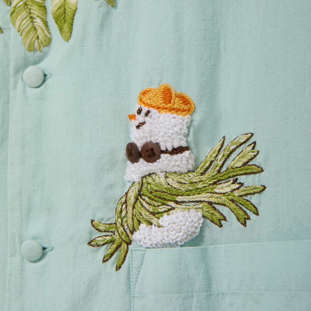Close-up of a light blue cabana shirt showing embroidered artwork of a small snowman wearing sunglasses and sitting on embroidered palm fronds above a front pocket.