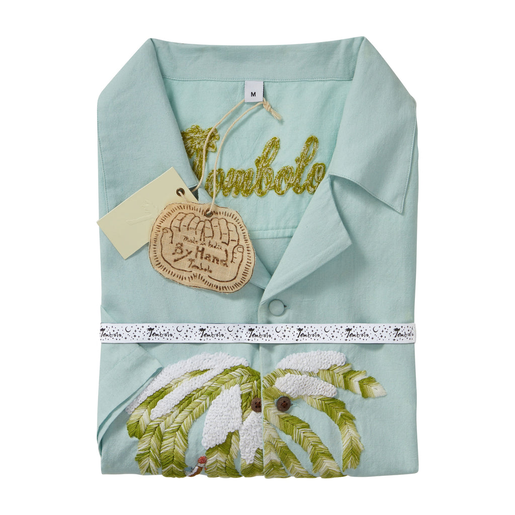 Folded light blue cabana shirt with embroidered snowy palm tree detail, shown with a chain-stitched “Tombolo” logo at the collar. The shirt is wrapped with branded ribbon and includes attached hangtags.